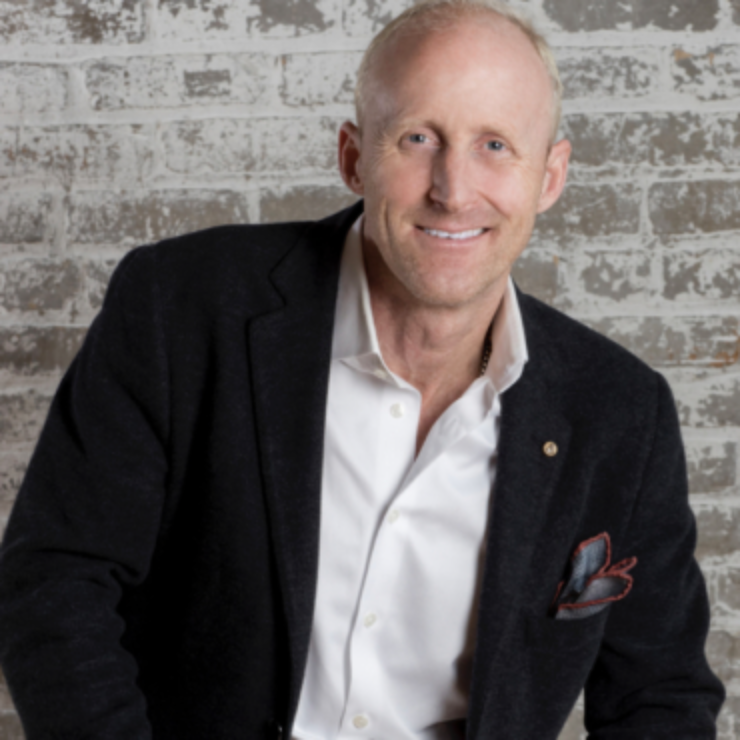 Smiling man in a black blazer and white shirt poses against a brick wall.